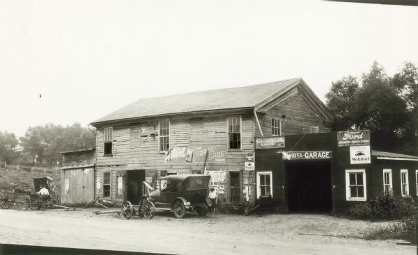 A mower and car parked in front of the dilapidated Mayne Hotel. Two boys are standing to the right of the car, and two men are talking near a wagon and buggy on the left. A horse is inside the old hotel, now identified as the Wiota Garage. There are signs advertising Mobiloil, Ford service and Goodrich tires on an addition at the side of the main building. Signs on the building advertise Velvet and Plow Boy chewing tobacco and the "Big White Fair" at Burlington.