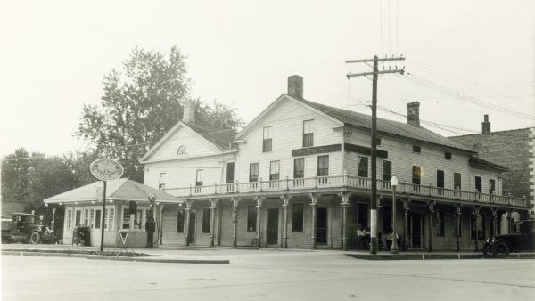 This substantial wooden hotel in the classical revival style has a large wing in the rear and another on the right. A wrap-around porch unifies the building; three men sit on the porch. There is a small filling station on the left identified as Hansen Oil Company with a truck parked nearby. There is also a car on the far right.
