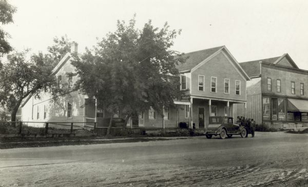 Two men stand in front of the wooden Central Hotel building while a third stands in the doorway. An automobile is parked in front. To the right is a second wooden commercial building with a large awning. The road is unpaved.