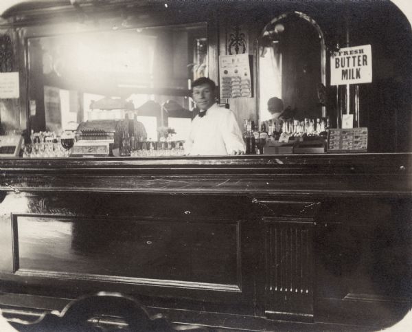 The bartender Art Gerth stands behind the bar of a tavern located at the corner of Twelfth and Chambers. A glass of beer stands on the bar.  Behind the bar is the cash register, glassware and bottles. The back bar has large mirrors. A sign advertises "Fresh Butter Milk."