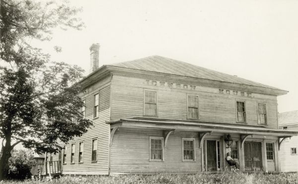 A man sits on the porch of the Johnson House hotel. The large wooden structure has a standing seam metal roof and tall chimney. "Johnson House" is painted on the front; large brackets support the porch roof.