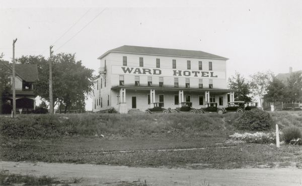 A three-story wooden structure with front porch identified as the Ward Hotel by a large sign on the front of the building. Several automobiles are parked diagonally in front. There are houses on either side.