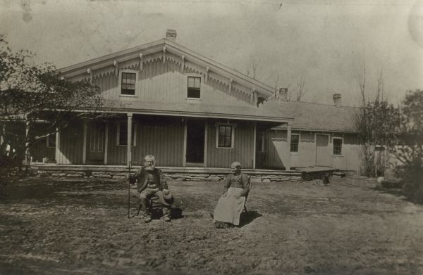 A copy of an older photograph of the William Plocker (Plokker) Tavern south of Fairwater. An unidentified couple sits in the front yard; the man holds a rake. The front (gable) end of the building has fancy eave brackets and a porch with stone foundation. There is a low wing to the right.