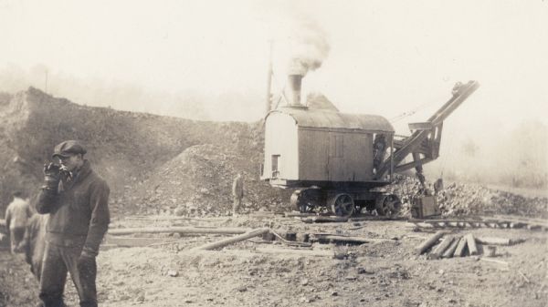 A workman smokes a cigarette in the foreground as a steam shovel loads iron ore at the Cahoon Mine.