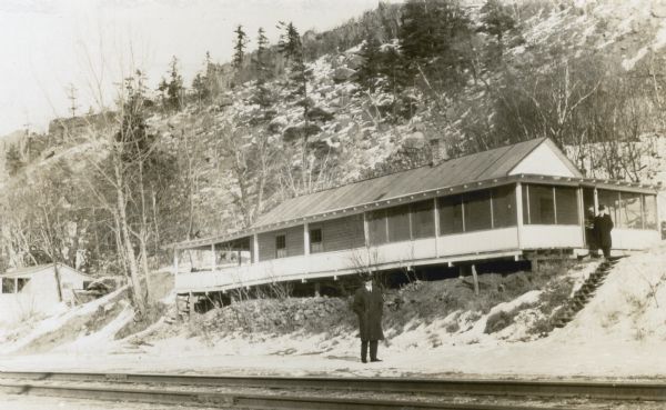 Highcliffe, a cottage with a large porch near the shore of Devil's Lake which was the meeting place of the Heart of the Hills Walking Club, founded by H.E. Cole. Two unidentified men pose for the photograph. There are railroad tracks in front of the cottage and the East Bluff rises behind. Snow is on the ground.