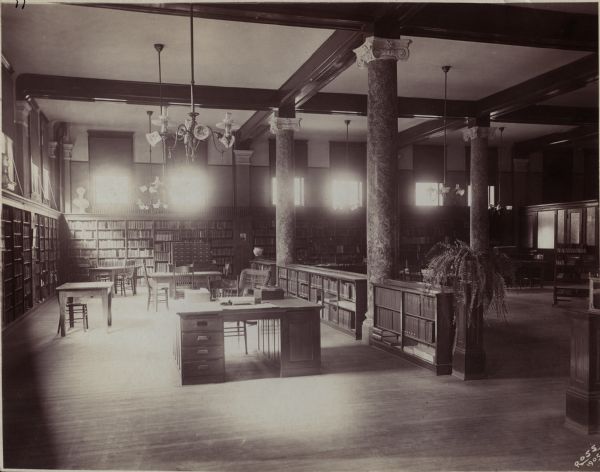 Interior view of the Appleton Free Public Library. There are reading tables, a librarian's desk, and bookshelves. Large light fixtures are hanging from the exposed beam ceiling, and there are three marble columns crowned with ionic capitals.