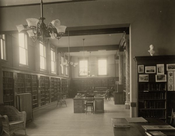 Interior view of the Appleton Free Public Library. There are reading tables, a librarian's desk, bookshelves, and a wicker chair. Two large light fixtures are hanging from the ceiling.