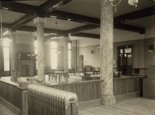 Interior view of the Appleton Free Public Library. In the large room are reading tables, fireplace, radiators, carved wood railings, and wicker furniture. Large light fixtures are hanging from the exposed beam ceiling, and there are four marble columns crowned with ionic capitals.
