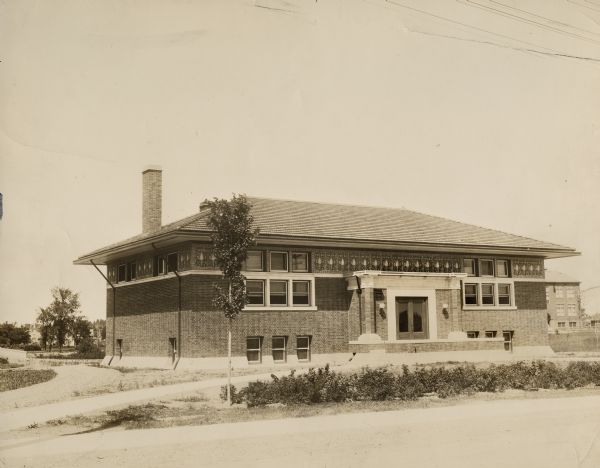 Exterior view of the Merrill Public Library. Above the main entrance it reads: "T.B. Scott Free Library." Reverse of the cardboard backing reads: "Carnegie bldg $17,500, 1910, Claude + Starck, arch. Madison, Style "Sullivanesque".<p>This note conflicts with information found in the "Tenth Biennial Report of the Wisconsin Free Library Commission, 1914," which states that the library was funded by a $15,000 gift from Andrew Carnegie and opened in 1911. Prominent elements of the photo: prairie style with Sullivanesque elements.<p>Additional notes from: "New Types of Small Library Buildings, 1913"
CONSTRUCTION - Concrete foundation; mission brick and ornamental frieze above tile roof; plate glass; oak finish first story; basement yellow pine; electric lights; steam heat. DIMENSIONS - 75'x38' with stack room 16'x26'. One story and basement. CAPACITY - 17,500 volumes; Floor plans included.</p>