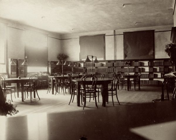 Interior view of the Beloit Public Library. There are reading tables with large lamps, bentwood chairs, and bookshelves throughout the room.