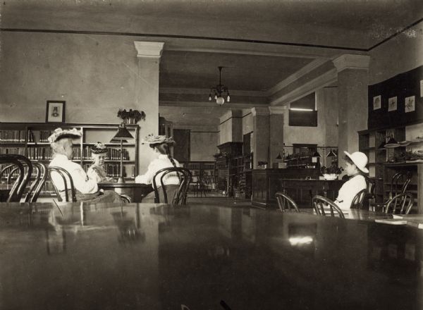 Interior view of the Beloit Public Library. The library opened in 1903 and Andrew Carnegie funded library construction with $25,000. Three women are seated around a table wearing hats, a young girl seated off to the side, bookshelves, a fireplace, and librarian's desk.