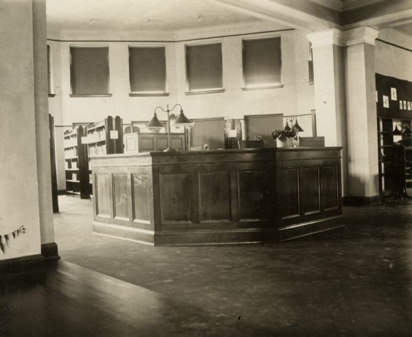 Interior view, perhaps from the entrance, of the Beloit Public Library. There is a large librarian's desk in the center, with freestanding bookshelves on both sides.