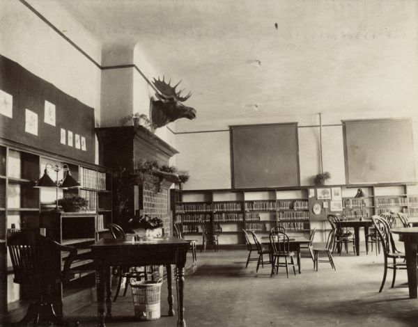 Interior view of the Beloit Public Library. This image is also available as a cyanotype. The reverse of the cyanotype version has a note: "Patton + Miller, Chicago, Arch., Carnegie bldg, 1903 $25,000." There are reading tables, chairs, bookshelves, a fireplace, and mounted moose head on the wall.