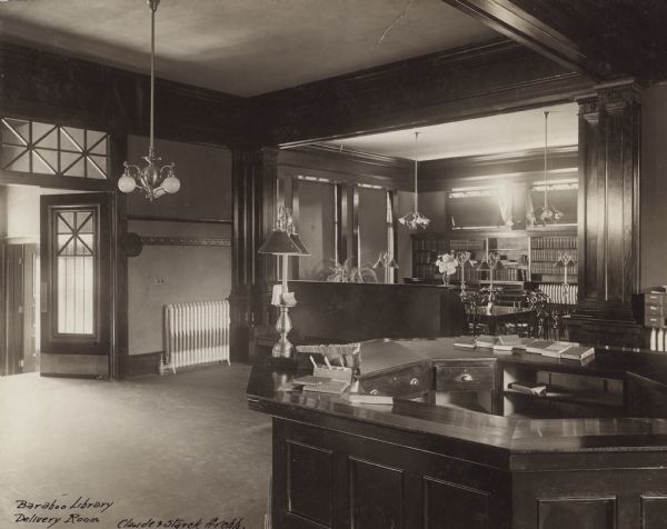 Interior view of the Baraboo Carnegie Free Library Delivery Room. Text at lower left: "Baraboo Library, Delivery Room, Claude & Starck Archts." There is a large librarian's desk, bookshelves, and a reading table.