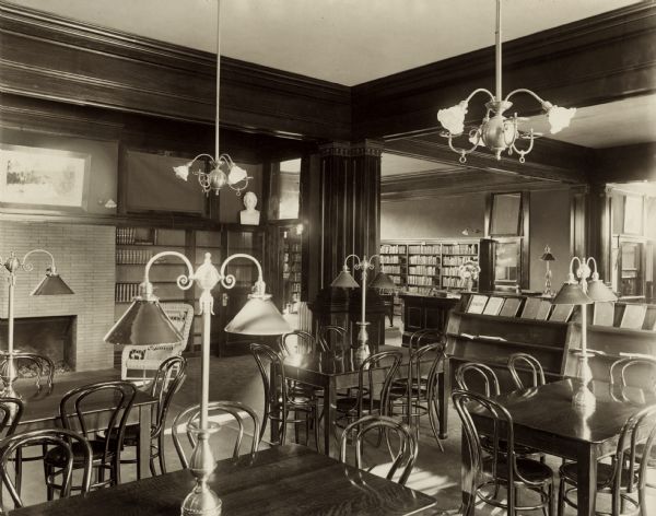 Interior view of the Baraboo Carnegie Free Library. There is a fireplace, bookshelves, display shelves, and reading tables with bentwood chairs. There are large lamps on the tables, and two light fixtures hanging from the ceiling.