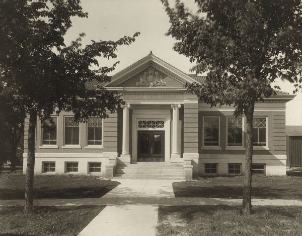 Exterior view of front entrance, framed by two trees, of the Baraboo Carnegie Free Library. Above the main entrance it reads: "Carnegie Free Library." On the reverse of the cardboard backing it reads: "Claude and Starck, Madison, 1903, cost $15,000."