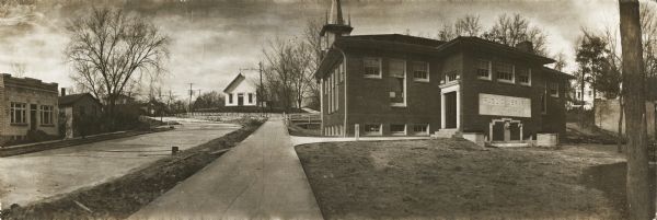 Panoramic picture postcard of the exterior of the Black River Falls Public Library. One side of the library has a large white stone on which is carved: "Public Library / Black River Falls." On the back of the cardboard backing are the words: "Black River Falls Lib. $10,000." The image includes a view of the curved street and several unidentified buildings. There is a steeple visible behind the library building.