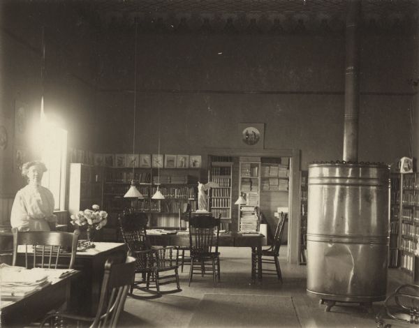 Interior view of the Black River Falls Public Library. In the large, open room a woman (librarian?) is standing on the left near a librarian's desk. A large cylindrical metal furnace with exhaust pipe is on the right, and the the background are bookshelves and a statue of Winged Victory.<p>Additional notes from Biennial Reports of the Free Library Commission, 1898-1936.<br>Mrs. Mary J. Gunn is named as the librarian of Black River Falls Library in the 2nd (1897-98) & 3rd (1899-1900) biennial reports of the Library Commission, during these years library was located in City Hall. In 1900 the public library moved to the school building. In Fall of 1914 construction was started on a Carnegie funded library building that opened in 1915. The 4th (1901-02) - 21th (1935-36) biennial reports of the Library Commission (all this author has access to) list Annie C. Wylie as librarian.</p>