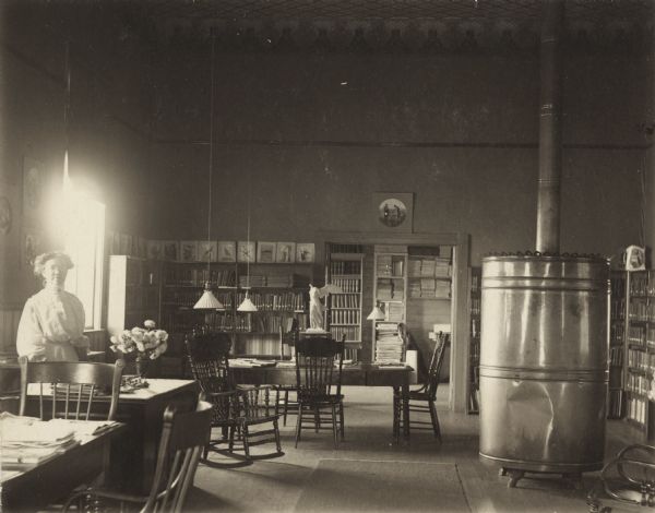 Interior view of the Black River Falls Public Library. In the large, open room a woman (librarian?) is standing on the left near a librarian's desk. A large cylindrical metal furnace with exhaust pipe is on the right, and the the background are bookshelves and a statue of Winged Victory.<p>Additional notes from Biennial Reports of the Free Library Commission, 1898-1936.<br>Mrs. Mary J. Gunn is named as the librarian of Black River Falls Library in the 2nd (1897-98) & 3rd (1899-1900) biennial reports of the Library Commission, during these years library was located in City Hall.  In 1900 the public library moved to the school building. In Fall of 1914 construction was started on a Carnegie funded library building that opened in 1915. The 4th (1901-02) - 21th (1935-36) biennial reports of the Library Commission (all this author has access to) list Annie C. Wylie as librarian.</p>