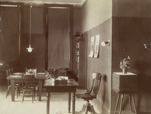 Interior view of the Brodhead Public Library. There is a small librarian's desk with a tray of cards and flowers in a vase. On the right is a nine-drawer card catalog on a small table, and in the background is a reading table with chairs, and one bookshelf. The tall windows have the shades drawn.