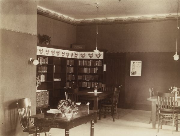 Interior view of the Brodhead Public Library. There is a poster on the wall advertising "The Debtor, a novel by May E. Wilkens Freeman." There are bookshelves in the corner of the room labeled "Fiction." In the foreground is a librarian's desk with two trays of cards and avase of fresh flowers, a nine-drawer card catalog is just behind a corner near two reading tables.