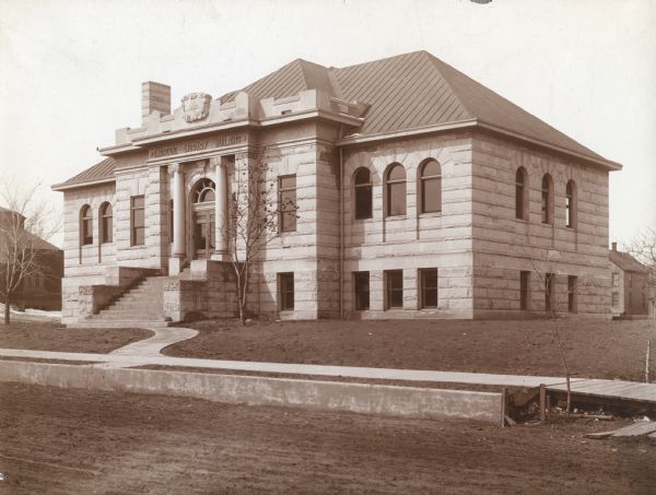 Exterior view from road of the Chippewa Falls Library. The library opened in 1903 and Andrew Carnegie funded the library with $10,000. Above the main entrance it reads: "Carnegie Library Building." There is a crest above the entrance that reads: "AD 1902." Two trees frame the curved walkway and flight of steps up to the entrance.