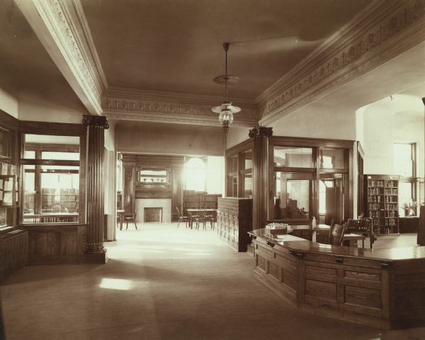 Interior view of the Chippewa Falls Library. There is a large open area with a librarian's desk with card trays on the right. A card catalog is in the space between two glassed-in offices, and a large room in the background has bookshelves, a fireplace, and reading tables.