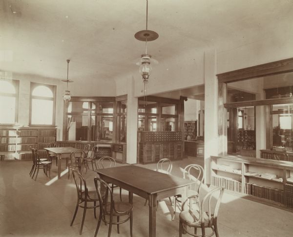 Interior view of the Chippewa Falls Library. In the foreground are reading tables and chairs. Bookshelves line the room. A large card catalog is in the space between two glass-walled offices, and the librarian's desk is in the space beyond.