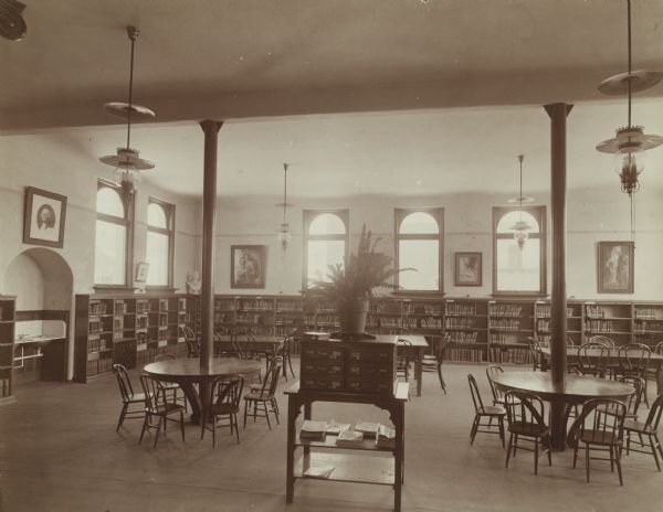 Interior view of the Chippewa Falls Library. In the center is a six drawer cabinet holding a potted plant, surrounded by small reading tables (children's tables?), and adult reading tables. Two of the round tables are built around columns. Bookshelves line the walls on which are hanging framed prints. (George Washington, Madonna and child). There is a sink in an alcove on the left.