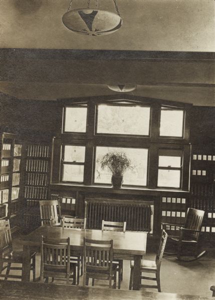 Interior view of the Columbus Library. There are reading tables and chairs, and bookshelves line the walls. A large window with a potted plant is in the center. Two lamps hang from the tall ceiling.