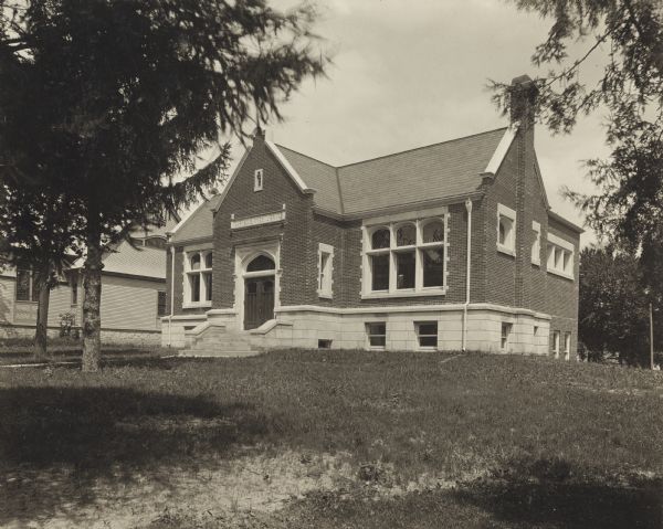 Exterior view from lawn of the Darlington Public Library. Above the main entrance it reads: "Carnegie Free Library." On the reverse of the photograph is reads: "Claude + Starck, Madison, arch., 1904, Carnegie Bldg., $10,000." Steps lead up to the entrance of the brick and stone building with a chimney on the right side.

Additional notes from "Small Library Buildings; A Collection of Plans Contributed by the League of Library Commissions, 1908."
COST - $10,117.73. Basement unfinished, cost of completion $600. 12.83c per cubic foot. Contract $9,297 (including book cases and loan desk). Heating plant $770, Lighting plant $135, Plumbing $200, Floor covering $203.10, Architect's fee $379.65, Furniture $168. CONSTRUCTION - Gothic architecture. Pressed brick and Bedford stone, with slate roof. Plate glass. Oak finish inside. Pine floors covered with cork carpet. Steam heat. Electric lights in ceiling and on each table. Natural light excellent from high windows at rear. Ventilated from windows and fireplace. Oak wall shelving 7'2" high. Low shelving with corticene bulletin above in children's room. DIMENSIONS - 60x48. Main Floor 12'6". high basement 10'. CAPACITY - 5,000 volumes, without use of floor cases. Seating capacity 20 each for reference and children's rooms, 32 for book room at rear.