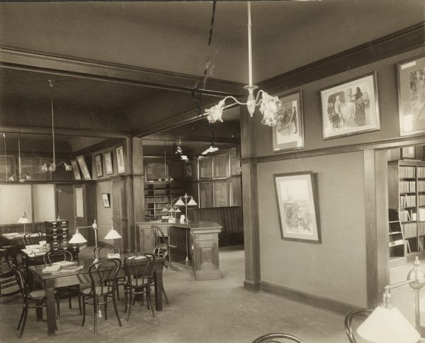 Interior view of the Darlington Public Library. In the foreground are tables and chairs, an eight drawer card catalog, and a lamp hanging from the ceiling. In the background is a librarian's desk, bookshelves, and a large wooden bench. There are framed prints in the middle of a wall that divides two rooms.