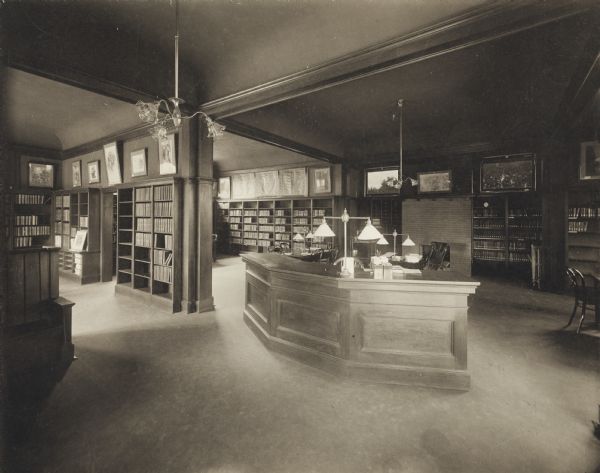 Interior view of the Darlington Public Library. In the foreground is a librarian's desk, and a bench on the left. In the background in two separate rooms are bookshelves, a fireplace, and framed prints on the walls.