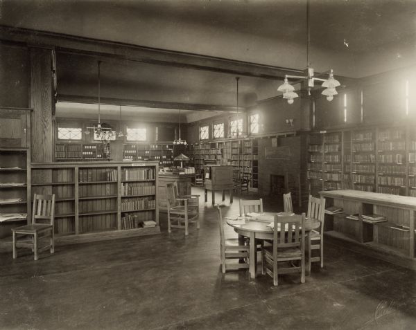 Interior view of the Aram Public Library; 404 East Walworth Avenue. The library was opened in 1908 and James Aram funded the library with a gift of $20,000. The lower right corner is stamped: "Curtiss, Delavan, Wis." In the foreground are children's table and chairs. Behind are  bookshelves, a fireplace, a free standing card catalog, and a librarian's desk.