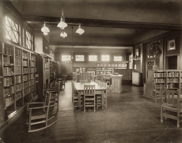 Interior view of the Aram Public Library; 404 East Walworth Avenue. The lower right corner is stamped: "Curtiss, Delavan, Wis." In the foreground is a reading table, and a rocking chair. Bookshelves line the walls, and there is a fireplace on the left wall. In the middle of the room is a free standing card catalog and a librarian's desk.
