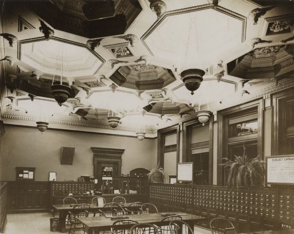 Interior view of the Dudgeon Library. An ornate coffered ceiling from which lamps hang from chains dominates the room. There are two large signs that read "Subject Catalogue" over the card catalogs. Card catalogs line the walls, and along the back wall in front of a doorway is a librarian's desk.