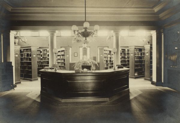 Interior view of the Eau Claire Public Library. Reverse of the photograph reads: "Carnegie bldg, 1904, Cost $40,000, Patton + Miller, Chicago, arch." In the center is a large librarian's desk, with a large chandelier above it. In the background are rows of bookshelves.