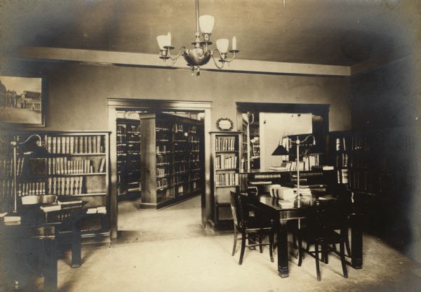 Interior view of the Eau Claire Public Library. There are tables with lamps, chairs, bookshelves, and a chandelier. Through a doorway is another room with bookshelves.
