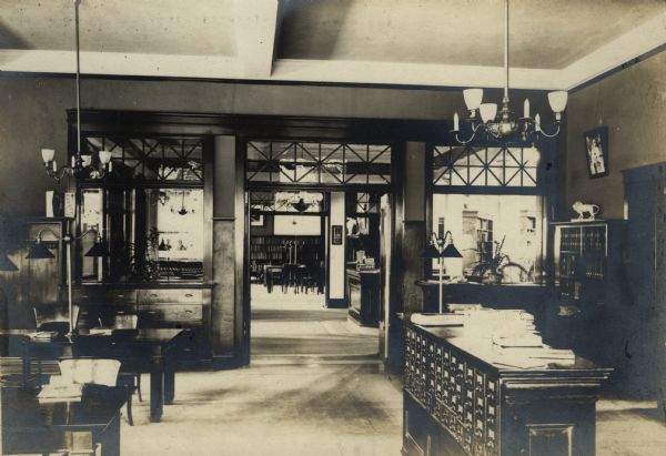 Interior view of the Eau Claire Public Library. Photograph is a long view through several rooms. The reverse of the photograph reads, "From children's room into the delivery room." In the foreground are reading tables and a card catalog, with light fixtures hanging from the tall ceiling.

Additional notes from "New Types of Small Library Buildings, 1913."
ARCHITECTS - Patton & Miller, Chicago. FUNDING SOURCE - Andrew Carnegie, $40,000. COST- $40,003.87. Contract $29,981.20 (including cork carpet and wall shelving). Tile roof $1,600. Gable ornaments $138. Steam pipes in gutters $42. Heating and ventilating $1,719, Electric wiring $5.22, Light fixtures $550, Plumbing and gas fitting $904., Shades $65, Decorations $810, Leaded glass $312, Architect's fees $1,884.66, Superintendence $384, Chairs $295, Catalogue drawers $69, Catalogue case, loan desk, tables and librarian's desk $728. Includes floor plans.