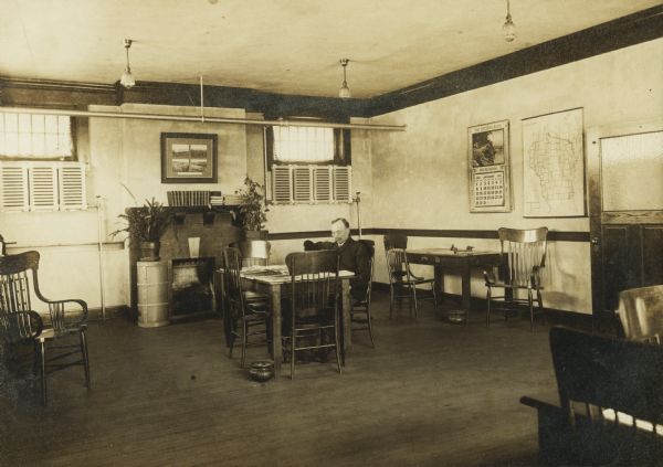 Interior view of the Elroy Public Library. The library opened in 1908 and was funded by Andrew Carnegie with $10,000. On the lower right the cardboard backing is stamped: "Fisk, Elroy, Wis." A man is seated at one of the reading table, and next to the reading tables are spittoons. There is a fireplace on the back wall, and on other walls are a map of Wisconsin and a Citizen Bank Calendar for January 1911.