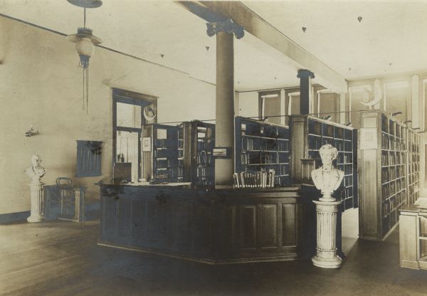 Interior view of the Janesville Public Library. There is large librarian's desk, with a bust of Abraham Lincoln to the right. Behind the desk are two columns and five rows of bookshelves. On top of the ends of two of the bookshelves are owls, perhaps stuffed, perched on a crescent moon.