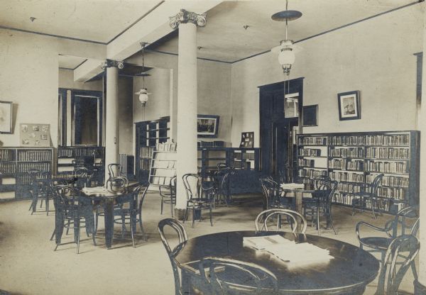 Interior view of the Janesville Public Library. Among the room are reading tables, bentwood chairs, light fixtures hanging from the tall ceiling, and two columns. Bookshelves line the walls.