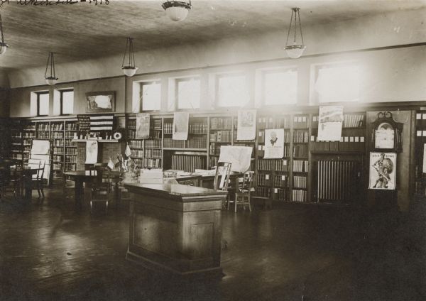 Interior view of the Fort Atkinson Public Library. The library was funded by H.E. Southwell with $10,000. War posters are prominently displayed and include "USA Bonds" and "Feed a Fighter." In the foreground is a librarian's desk, and behind it are reading tables,  bookshelves along the walls, war posters, a fireplace, grandfather clock, and a newspaper rack.