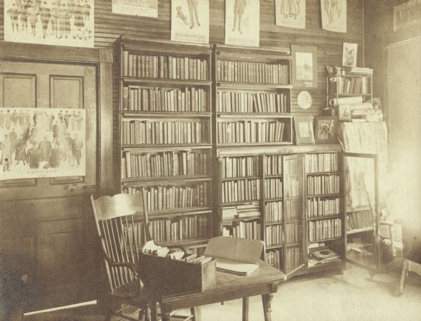 Interior view of the Genoa Junction Free Public Library. Reverse of cardboard mounting reads: "Genoa Junction Free Public Library, Genoa Junction, Wisconsin, Mrs. Carrie L. Manor Librarian. October 7, 1905." The library appears to be located in a dry goods store. On the back wall near a door are posters of men's fashions displayed around bookshelves. A small table with a small card catalog box and open register.