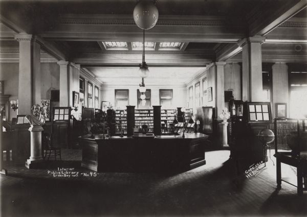 Interior view of the Green Bay Public Library. Notes on photograph read: "Interior, Public Library, Green Bay Wis. - No. 45" and "Copr. 1910, H.E. Bethe." Note on reverse of mounting reads: "Carnegie bldg. 1903, Cost $12,000, H.A. Foeller, Green Bay, arch." In the foreground is a large librarian's desk, surrounded by a card catalog, several busts on pedestals, globe, and a glass display case. In the back of the room tables, chairs, and bookshelves along the walls. Columns support the tall ceiling, and there is a skylight above the librarian's desk.