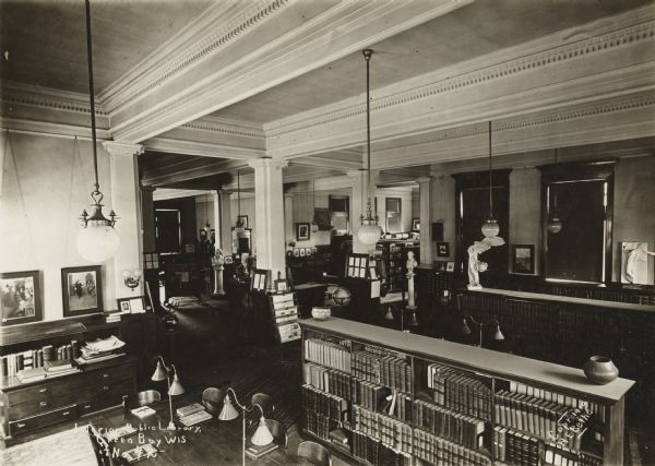 Elevated interior view of the Green Bay Public Library. The library was built in 1903 at a cost of $12,000 by H.A. Foeller, Architects. Notes on photograph read: "Interior, Public Library, Green Bay Wis. - No. 49" and "Copr. 1910, H.E. Bethe." Note on reverse of mounting reads: "Corner of Reference Room." There are reading tables with lamps, bookshelves, a card catalog, a newspaper rack, busts on pedestals, and a statue of Winged Victory.