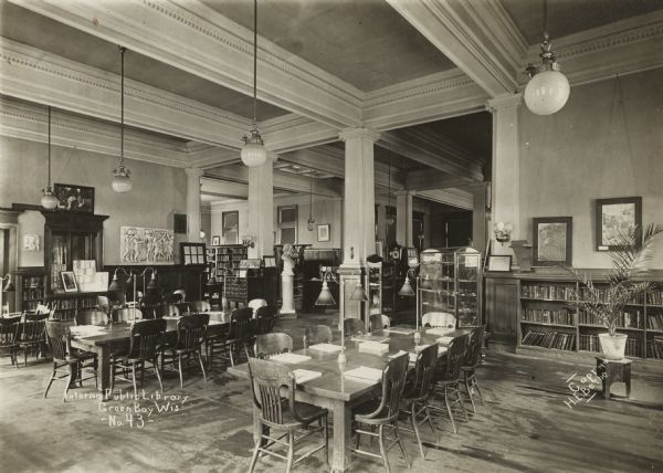 Interior view of the Green Bay Public Library. The library was built in 1903 at a cost of $12,000 by H.A. Foeller, Architects. Note on photograph reads: "Interior, Public Library, Green Bay Wis. - No. 43" and "Copr. 1910, H.E. Bethe." Note on reverse of mounting reads: "Children's Room." In the foreground are reading tables with chairs, bookshelves along the wall, a card catalog, busts on pedestals, a bas relief on a wall, and glass display cases.