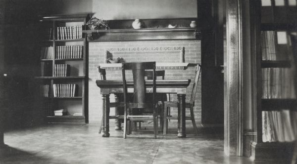 Interior view of the Hudson Public Library. The library opened in 1904 and was funded by Andrew Carnegie with a gift of $12,000. A reading table with high-backed chairs sits in front of a fireplace, with bookshelves along a wall on the left.