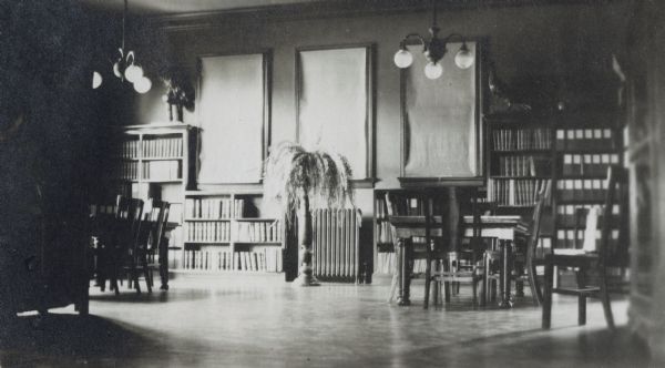 Interior view of the Hudson Public Library. The library opened in 1904 and was funded by Andrew Carnegie with a gift of $12,000. In the room are reading tables, bookshelves along the wall, a large fern on a plant stand, and a stuffed bird.