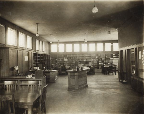 Interior view of the Jefferson Public Library. The library opened in 1910 and was funded by Andrew Carnegie with a gift of $10.000. Throughout the room are reading tables and chairs, a librarian's desk, and bookshelves. Windows line the room just below the ceiling above the bookshelves.<p>Additional notes from "New Types of Small Library Buildings, 1913."
ARCHITECTS - Claude & Starck, Madison, Wis. COST - $9,500. FUNDING SOURCE - Andrew Carnegie, $10,000. CONSTRUCTION - Original style. Stone basement; mission brick above stucco gables; slate roof; plate and art glass; oak finish first story; basement yellow pine; electric lights; steam heat. DIMENSIONS - 60'x34'. CAPACITY - 6,000 volumes. Identical floor plan to Reedsburg Public Library. Floor plans included.</p>